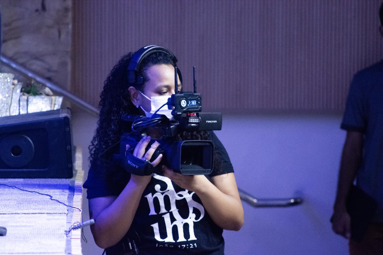 Letícia operando câmera em transmissão ao vivo, com fone e iluminação de palco
